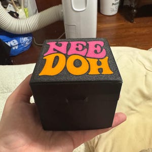 Puede incluir: Una caja cuadrada negra con superficie texturizada que presenta el texto NEE DOH impreso en la tapa superior. La palabra NEE es de color rosa y la palabra DOH es de color naranja. La caja tiene un panel frontal negro sólido y una tapa simple.