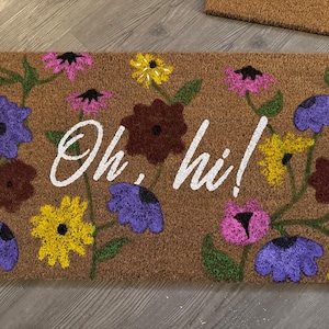 Puede incluir: Felpudo rectangular de fibra de coco marrón, adornado con diseños florales coloridos en morado, amarillo, rosa y marrón. Las palabras "Oh, hi!" están escritas en blanco sobre la alfombra, creando una entrada acogedora.