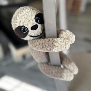 May include: A plush sloth toy made of light brown textured yarn, featuring dark brown patches around its blue eyes, a black embroidered nose, and a stitched smile. The sloth has long arms wrapped around a vertical gray post.