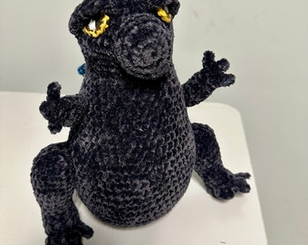 Puede incluir: Un juguete de peluche de crochet negro con forma de dinosaurio con ojos amarillos. El juguete está sentado sobre una superficie blanca.
