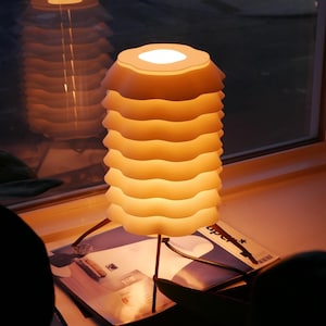 May include: A modern, layered lamp with a warm, inviting glow. The lamp features a stacked, wavy design in a soft, off-white color, supported by slender, dark legs. It sits on a table with reading material, near a window.