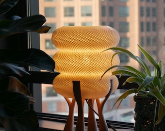 Op de afbeelding: Een tafellamp met drie gestructureerde, gebroken witte, donutvormige kappen die een warm licht uitstralen. De lamp wordt ondersteund door vier donkerbruine, slanke, vertakte poten die omhoog reiken om de basis van de lamp te ondersteunen.