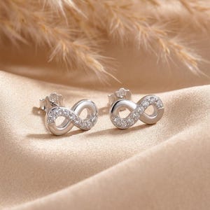 May include: A pair of silver stud earrings shaped like infinity symbols. Each earring features a polished silver finish on one side and a row of small, clear, sparkling stones embedded along the other side of the infinity loop design.