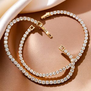 May include: Two gold-tone tennis bracelets with clear cubic zirconia stones. The bracelets are layered on top of each other.