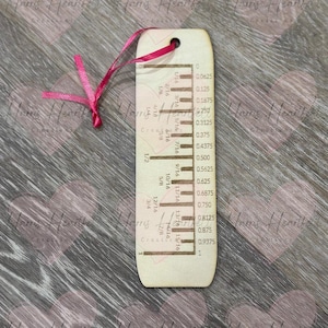 May include: A wooden bookmark with a ruler design, featuring measurements in inches and fractions. It has a pink ribbon attached at the top. The bookmark is light-colored with dark brown markings.