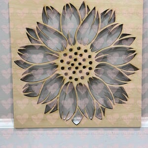 May include: A wooden sunflower wall art piece. The sunflower is laser cut with multiple layers, creating a 3D effect. The petals are light gray, and the center is dark brown with small, round details. The background is a light wood color.