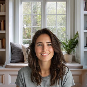 May include: A woman with long brown hair smiles in front of a window. Bookshelves filled with books are on either side of the window. Plants in pots sit on the windowsill and floor. The woman wears a grey t-shirt.