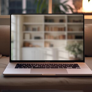 May include: A silver laptop computer with a black keyboard and trackpad is open on a light brown couch. The laptop screen is displaying a blurred image of a bookshelf.
