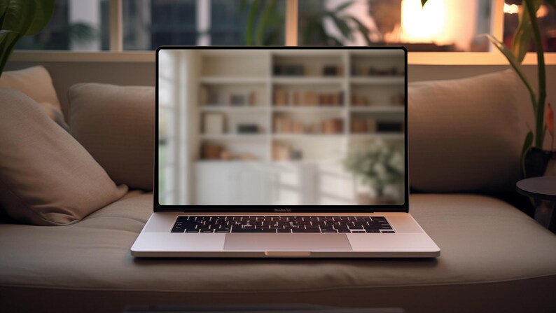 May include: A silver laptop computer with a black keyboard and trackpad is open on a light brown couch. The laptop screen is displaying a blurred image of a bookshelf.