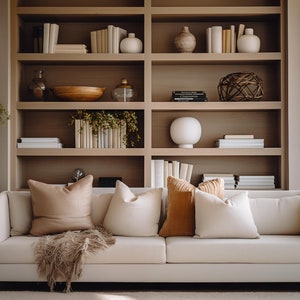 May include: A white sofa with a brown throw blanket and several pillows in a living room with a large bookshelf filled with books and decorative objects. The room has large windows with a view of trees outside.