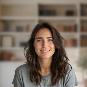 May include: A woman with long brown hair smiles at the camera. She is wearing a gray t-shirt and is standing in front of a bookshelf.