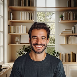 May include: A man with a beard and a dark blue shirt smiles at the camera in a room with a bookshelf and a window.