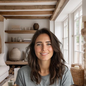 May include: A woman smiles in a cozy living room with exposed wooden beams and a stone fireplace. A white sofa with striped pillows sits near a window. Shelves display pottery and books. The room has a warm, inviting atmosphere.