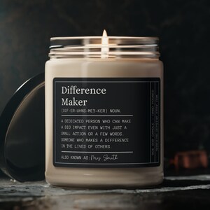 May include: A beige candle with a black label that reads "Difference Maker (DEF-ER-UHNS-MEY-KER) NOUN. A dedicated person who can make a big impact even with just a small action or a few words. Someone who makes a difference in the lives of others. ALSO KNOWN AS: Mrs. Smith"