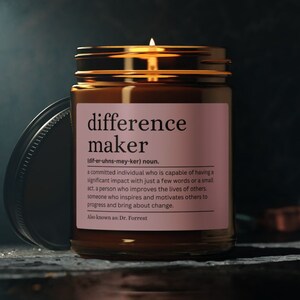 May include: A brown glass candle jar with a pink label that reads "difference maker" and a definition of the word. The candle is lit and the flame is visible.