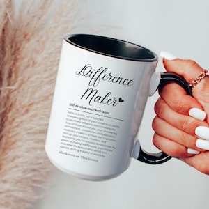May include: White ceramic mug with a black handle and black rim. The mug has a black and white graphic with the text "Difference Maker" and a definition of the term.