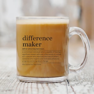 May include: A clear glass mug with a brown liquid inside. The mug has a black text graphic that reads "difference maker (dif-er-uhns-mey-ker) noun. a committed individual who is capable of having a significant impact with just a few words or a small act. a person who improves the lives of others. someone who inspires and motivates others to progress and bring about change. Also known as: Tara Foote"
