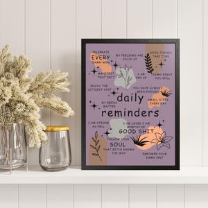 May include: A framed print with the words "daily reminders" in white text on a purple background. The print includes phrases like "Celebrate Every Damn Win" and "Follow Your Soul". Decorative floral and star elements are included.