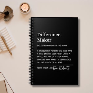 May include: Black and white notebook with a spiral binding. The cover has the text "Difference Maker" with the phonetic spelling "(DIF-ER-UHNS-MEY-KER)" and the definition "A dedicated person who can make a big impact even with just a small action or a few words. Someone who makes a difference in the lives of others. Also known as: Dr. Roberts"