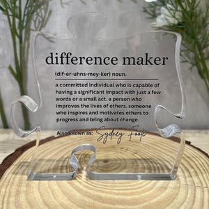 May include: A clear acrylic puzzle piece with the definition of a "difference maker" printed on it. The definition reads: "a committed individual who is capable of having a significant impact with just a few words or a small act. A person who improves the lives of others. Someone who inspires and motivates others to progress and bring about change." The text "Also known as: Sydney Foote" is printed below the definition.