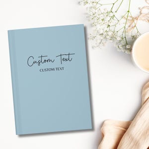 May include: A light blue notebook with the text "Custom Text" written in black cursive script. The text "CUSTOM TEXT" is written in all caps below the cursive text.