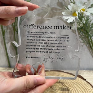 May include: A clear acrylic puzzle piece shaped like a jigsaw piece with the definition of "difference maker" printed on it. The definition reads: "(dif-er-uhns-mey-ker) noun. A committed individual who is capable of having a significant impact with just a few words or a small act. A person who improves the lives of others. Someone who inspires and motivates others to progress and bring about change. Also known as: Sydney Foste"