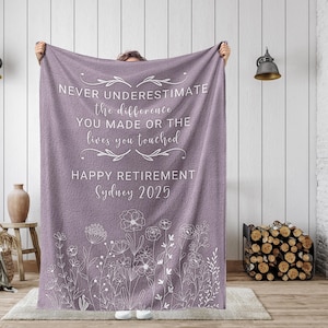 May include: Purple fleece blanket with white text that reads "NEVER UNDERESTIMATE the difference YOU MADE OR THE lives you touched HAPPY RETIREMENT Sydney 2025". The blanket has a white floral design at the bottom.
