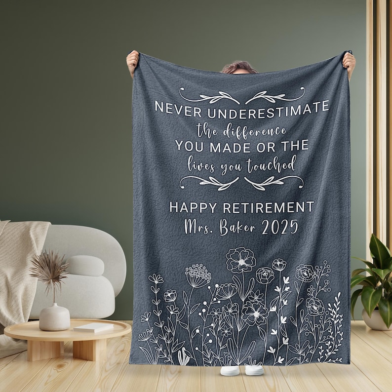 May include: A gray fleece blanket with white text that reads "Never underestimate the difference you made or the lives you touched. Happy Retirement Mrs. Baker 2025". The blanket has a white floral design at the bottom.