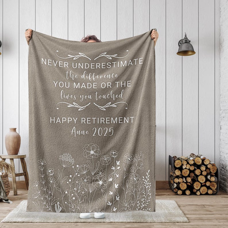 May include: A brown fleece blanket with white text that reads "NEVER UNDERESTIMATE the difference YOU MADE OR THE lives you touched HAPPY RETIREMENT Anne 2025". The blanket has a white floral design at the bottom.