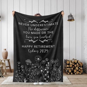 May include: Black fleece blanket with white text that reads "Never underestimate the difference you made or the lives you touched. Happy Retirement Sydney 2025". The blanket has a white floral design at the bottom.