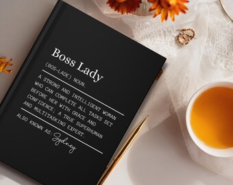 May include: A black notebook with the words "Boss Lady" in white. The definition of "Boss Lady" is printed below, along with the name "Sydney". A gold pen, a cup of tea, and flowers are in the background.