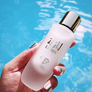 Peut inclure: Une bouteille en verre transparente avec un bouchon argenté et doré. La bouteille est étiquetée avec le mot "Laiqa" en arabe et en anglais. La bouteille est tenue par une main avec un vernis à ongles argenté.