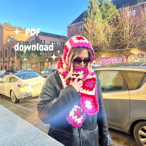 May include: A person wearing a pink and white crocheted hooded scarf with a white pom pom on top. The scarf is made of granny squares and has a flower design. The person is wearing a black leather jacket and is standing in front of a building.