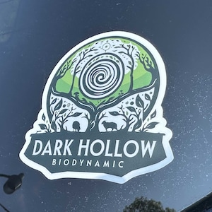 May include: A sticker with a nature-themed design. The sticker features a green and white illustration of trees, a spiral, and two animals. The text "DARK HOLLOW BIODYNAMIC" is displayed in white and green.