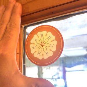 Puede incluir: Una pegatina redonda con un diseño floral. La pegatina presenta una flor color crema con un centro rojo y contornos negros, sobre un fondo naranja. La pegatina está colocada en una ventana.