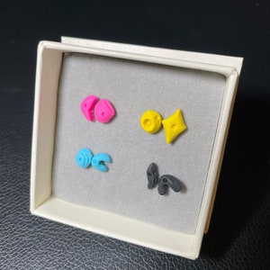 May include: A set of four colorful polymer clay earrings in a white box. The earrings are shaped like geometric figures, including a heart, a diamond, a circle, and a pair of abstract shapes. The earrings are pink, yellow, blue, and black.