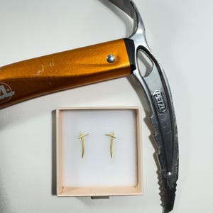 Op de afbeelding: Een oranje en zilveren ijsbijl naast een klein doosje met twee goudkleurige oorbellen. De oorbellen hebben een minimalistisch ontwerp, dat lijkt op een kruis of een gestileerde letter T.