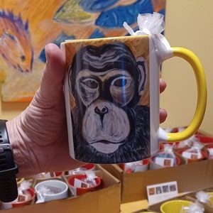 Può includere: Una tazza in ceramica bianca con un manico giallo presenta un'illustrazione dipinta a mano del viso di uno scimpanzé. La tazza ha un bordo giallo e una fascia gialla attorno al manico. Il viso dello scimpanzé è dipinto in tonalità di marrone, nero e bianco. La tazza è tenuta da una mano che indossa un orologio nero.