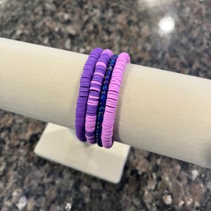 May include: Four bracelets in shades of purple and blue are displayed on a white stand. The bracelets are made of small, flat, round beads. The colors include dark purple, light purple, and a dark blue with glitter.