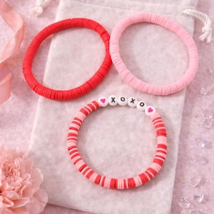 May include: Three colorful bracelets on a white pouch. One is solid red, another is pink, and the third is red and pink striped with XOXO beads. The bracelets are made of small, flat, round beads. The background is a soft pink.