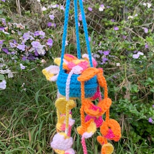 May include: A crocheted hanging mobile with a blue pot and colorful flower-shaped crocheted pieces. The mobile is hanging from a blue cord.