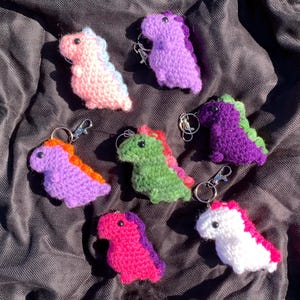 May include: Seven crocheted dinosaur keychains in various colors, including purple, pink, green, and white. Each keychain features a small, detailed dinosaur with a key ring attached.