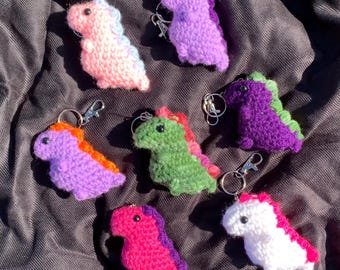 May include: Seven crocheted dinosaur keychains in various colors, including purple, pink, green, and white. Each keychain features a small, detailed dinosaur with a key ring attached.