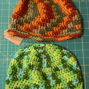 May include: Two handmade crochet beanies. The top beanie features a mix of orange, brown, and green yarn. The bottom beanie is green with lighter green and blue accents. Both beanies are on a green cutting mat.