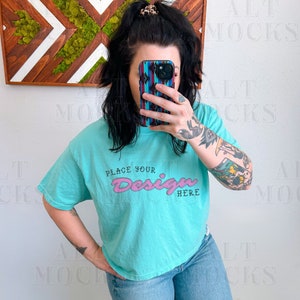 May include: A turquoise blue cropped t-shirt with short sleeves and the text "Place Your Design Here" in pink lettering. The shirt is being worn by a person with tattoos on their arms.