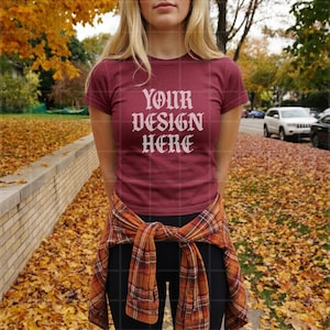 Può includere: Una t-shirt a maniche corte color bordeaux con la scritta bianca YOUR DESIGN HERE stampata sul petto. Una camicia di flanella a quadri nei toni dell'arancione, marrone e nero è legata in vita sopra leggings neri, su uno sfondo autunnale.