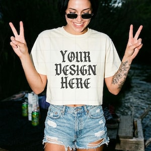 Könnte beinhalten: Ein cremefarbenes kurzärmeliges bauchfreies Oberteil mit dem schwarzen Text "YOUR DESIGN HERE" in einer gotischen Schriftart. Das Shirt wird mit einer hellblauen Jeansshorts kombiniert, die abgenutzte Ränder und sichtbare Risse aufweist.