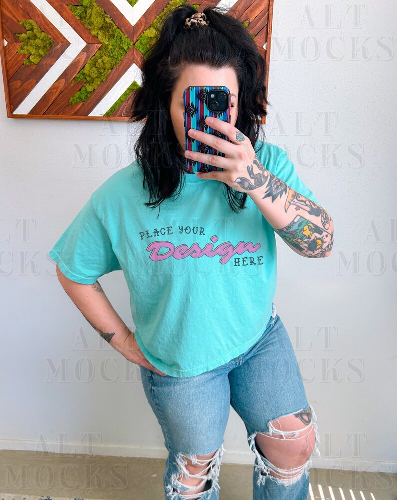 May include: A turquoise blue cropped t-shirt with short sleeves and the text "Place Your Design Here" in pink lettering. The shirt is being worn by a person with tattoos on their arms.