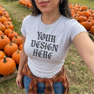 May include: A light gray short-sleeve t-shirt featuring the black text "YOUR DESIGN HERE" in a gothic-style font. The shirt is worn with a brown and orange plaid flannel shirt tied around the waist and blue denim jeans.