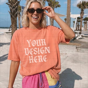 May include: A coral-colored t-shirt with the text "YOUR DESIGN HERE" in a white, ornate font. The shirt is paired with pink, yellow, and blue shorts. The person is wearing sunglasses and smiling.